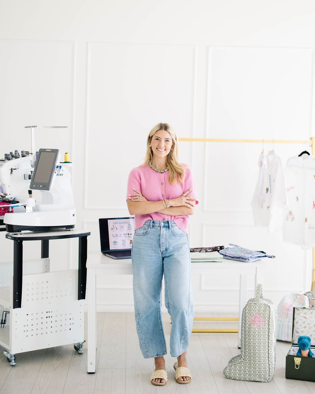 Madison in the studio with her embroidery machine, a rack of embroidered shirts and monogrammed bags