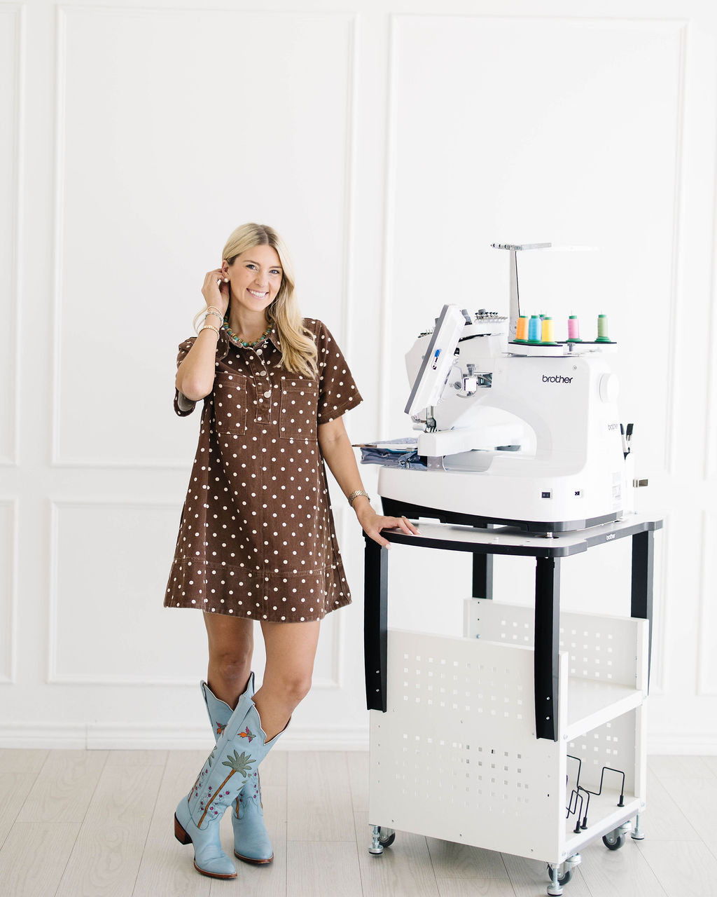 A woman stands next to a white Brother embroidery machine mounted on a white and black rolling cart in a bright, white-paneled room. She is wearing a brown short-sleeve polka-dot dress and light blue western boots.